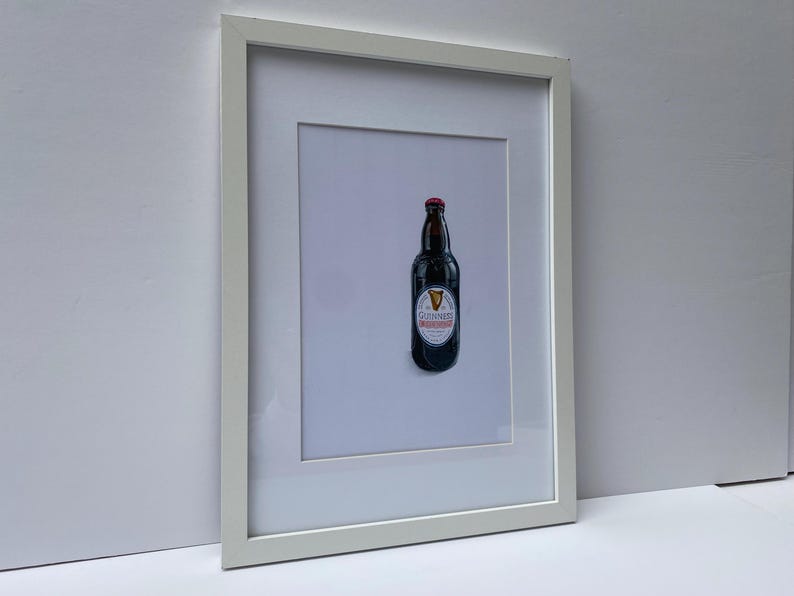 May include: Framed print featuring a dark brown Guinness beer bottle with a red cap and a white and red label. The framed artwork is displayed against a white background, encased in a white frame.