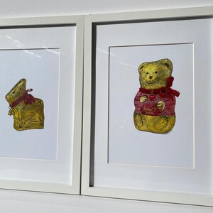 Puede incluir: Dos impresiones enmarcadas con figuras de chocolate envueltas en papel dorado. Una impresi&oacute;n muestra un conejito envuelto con una cinta roja y una campana. La otra impresi&oacute;n representa un oso de peluche con un su&eacute;ter rojo con un coraz&oacute;n.