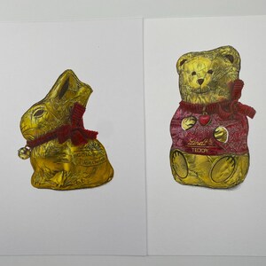Puede incluir: Dos figuras de chocolate envueltas en papel dorado: un conejito dorado con lazo y campana rojos, y un osito dorado con un su&eacute;ter rojo con un coraz&oacute;n. El oso tiene el texto "Lindt TEDDY".