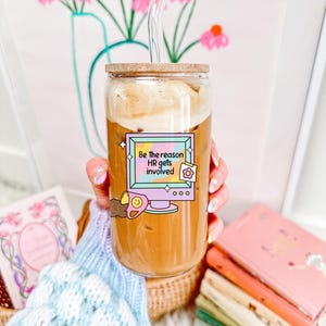 Puede incluir: Un vaso de vidrio transparente con tapa de madera y pajita de vidrio, lleno de una bebida de caf&eacute;. El vaso tiene una pegatina que dice "Be the reason HR gets involved". El fondo incluye una impresi&oacute;n floral y libros.