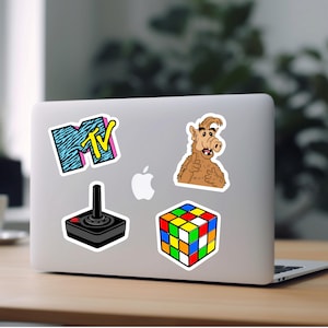 May include: A laptop computer with five stickers on it. The stickers are of a Rubik's Cube, a joystick, a cartoon character, and the MTV logo.