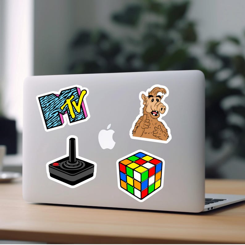 May include: A laptop computer with five stickers on it. The stickers are of a Rubik's Cube, a joystick, a cartoon character, and the MTV logo.