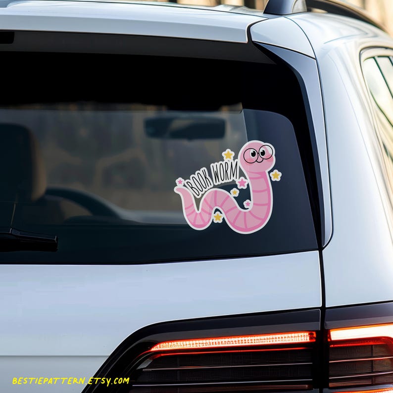 May include: A pink "Book Worm" sticker featuring a cartoon worm with large eyes and stars. The worm is in a curved shape, with the words "Book Worm" in a script font. The sticker is on a car window.