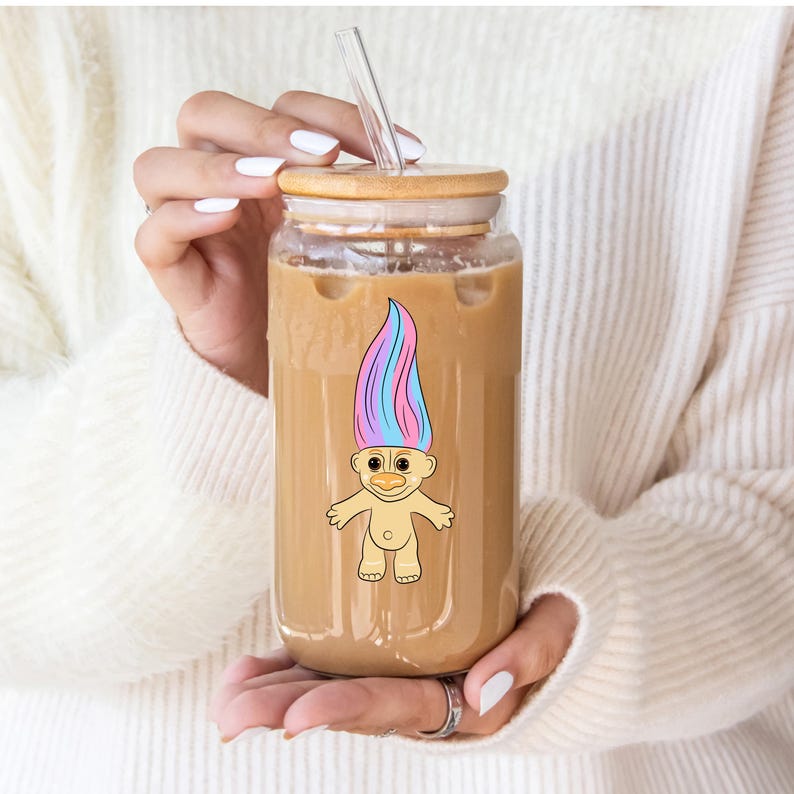 May include: A clear glass tumbler with a wooden lid and a straw. The tumbler is filled with iced coffee and has a sticker of a cartoon troll with blue and pink hair.