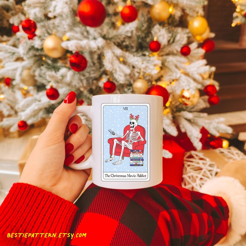 May include: White ceramic mug with a Christmas-themed design. The design features a skeleton wearing a Santa hat and sitting in a red armchair, watching TV. The text on the mug reads "The Christmas Movie Addict".