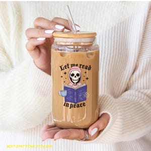 May include: A clear glass tumbler with a bamboo lid and a clear straw. The tumbler features a graphic of a skeleton reading a book with the text "Let me read in peace". The tumbler is filled with a light brown liquid.