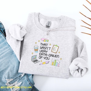 May include: A light gray sweatshirt with the phrase "THAT WASN'T VERY DATA-DRIVEN OF YOU" printed on the front. The design includes a pie chart, coffee cup, laptop, pen, clipboard, and bar graph. The sweatshirt is paired with blue jeans.