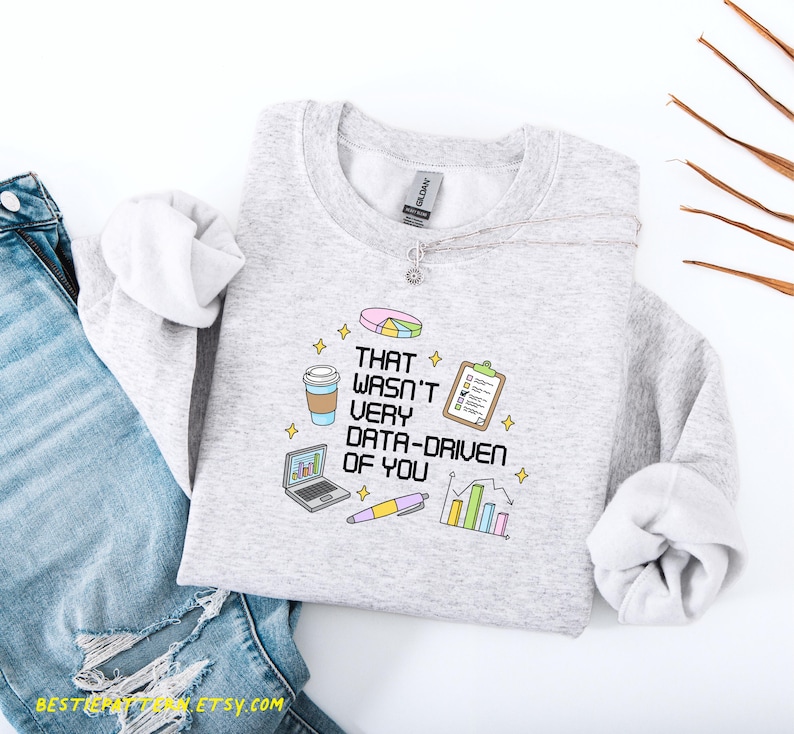 May include: A light gray sweatshirt with the phrase "THAT WASN'T VERY DATA-DRIVEN OF YOU" printed on the front. The design includes a pie chart, coffee cup, laptop, pen, clipboard, and bar graph. The sweatshirt is paired with blue jeans.