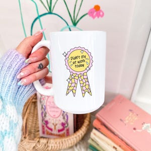 Puede incluir: Taza de cer&aacute;mica blanca con una ilustraci&oacute;n colorida de una cinta y el texto "Didn't cry at work today." La taza est&aacute; sostenida en una mano. Libros y una cesta se ven en el fondo.