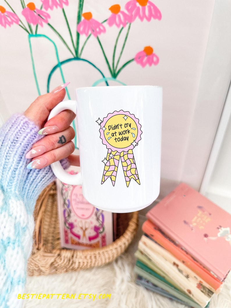 Puede incluir: Taza de cer&aacute;mica blanca con una ilustraci&oacute;n colorida de una cinta y el texto "Didn't cry at work today." La taza est&aacute; sostenida en una mano. Libros y una cesta se ven en el fondo.