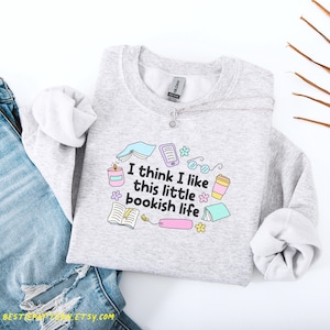 以下が含まれることがあります： 「I think I like this little bookish life」の文字と本をテーマにしたグラフィックがプリントされたライトグレーのスウェットシャツ。デザインには、開いた本、メガネ、キャンドル、コーヒーカップが含まれています。スウェットシャツはブルージーンズと組み合わせています。