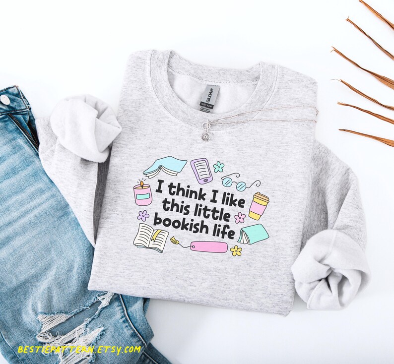 以下が含まれることがあります： 「I think I like this little bookish life」の文字と本をテーマにしたグラフィックがプリントされたライトグレーのスウェットシャツ。デザインには、開いた本、メガネ、キャンドル、コーヒーカップが含まれています。スウェットシャツはブルージーンズと組み合わせています。