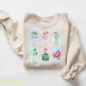 May include: A beige sweatshirt with a graphic print of skeletons dressed in Christmas attire. The skeletons are holding gifts, drinking champagne, and wearing Santa hats. The text on the sweatshirt reads "The Romantic", "The Gifter", "The Strangely", "The Drinker", "The Christmas Lover", and "The Christmas Movie Addict".