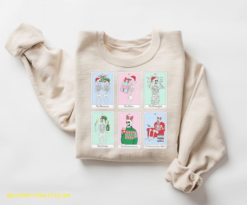 May include: A beige sweatshirt with a graphic print of skeletons dressed in Christmas attire. The skeletons are holding gifts, drinking champagne, and wearing Santa hats. The text on the sweatshirt reads "The Romantic", "The Gifter", "The Strangely", "The Drinker", "The Christmas Lover", and "The Christmas Movie Addict".