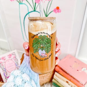 May include: A clear glass tumbler with a bamboo lid and a glass straw, filled with a coffee drink. The tumbler features a green illustration of a plant and the text "I strive to be as strong as the neglected office plant."