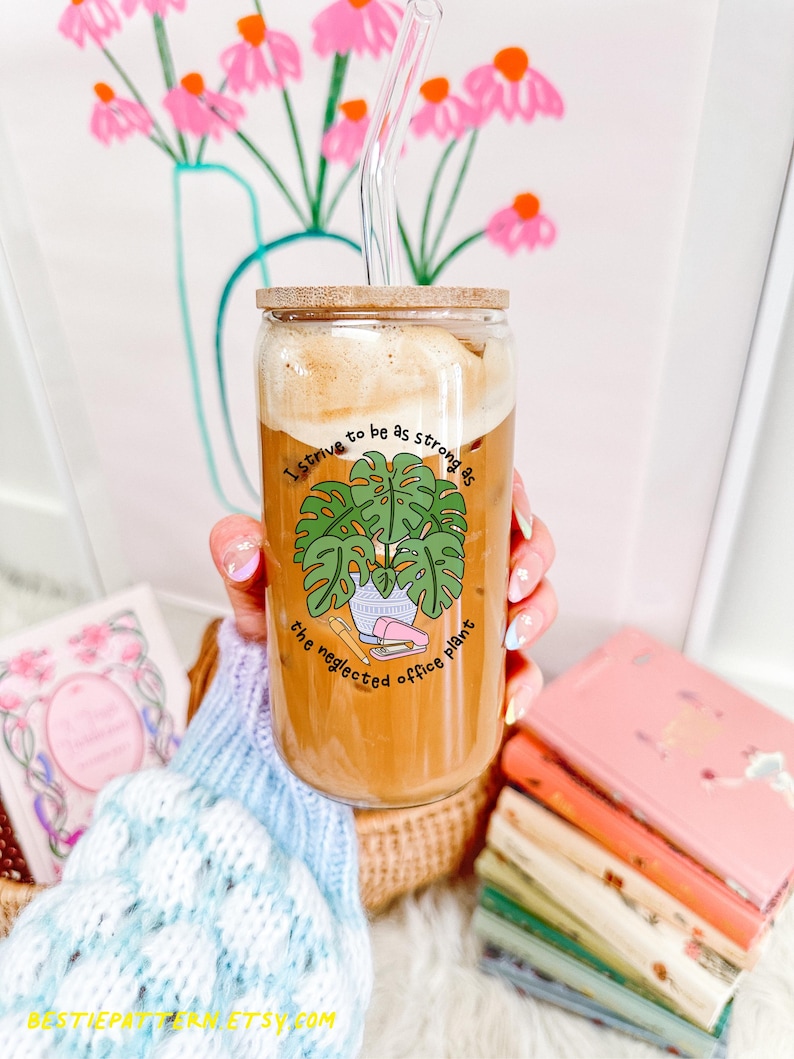 May include: A clear glass tumbler with a bamboo lid and a glass straw, filled with a coffee drink. The tumbler features a green illustration of a plant and the text "I strive to be as strong as the neglected office plant."