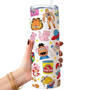 May include: A white tumbler with a straw, featuring a colourful pattern of 80s cartoon characters, including Garfield, He-Man, Care Bears, Strawberry Shortcake, and Play-Doh.