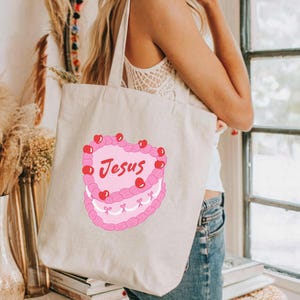 Puede incluir: Una bolsa de lona blanca con un dise&ntilde;o de pastel rosa en forma de coraz&oacute;n. El pastel tiene cerezas rojas y la palabra "Jes&uacute;s" escrita en &eacute;l.