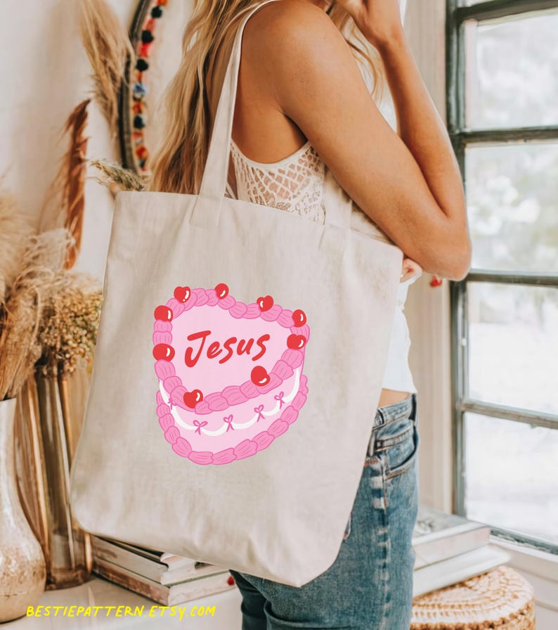 Puede incluir: Una bolsa de lona blanca con un dise&ntilde;o de pastel rosa en forma de coraz&oacute;n. El pastel tiene cerezas rojas y la palabra "Jes&uacute;s" escrita en &eacute;l.