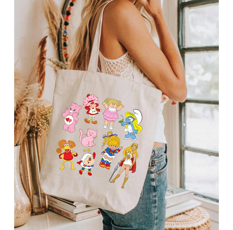 Puede incluir: Una bolsa de lona beige con un estampado colorido de personajes de dibujos animados de programas populares de los a&ntilde;os 80, como Care Bears, Strawberry Shortcake, los Pitufos y She-Ra.