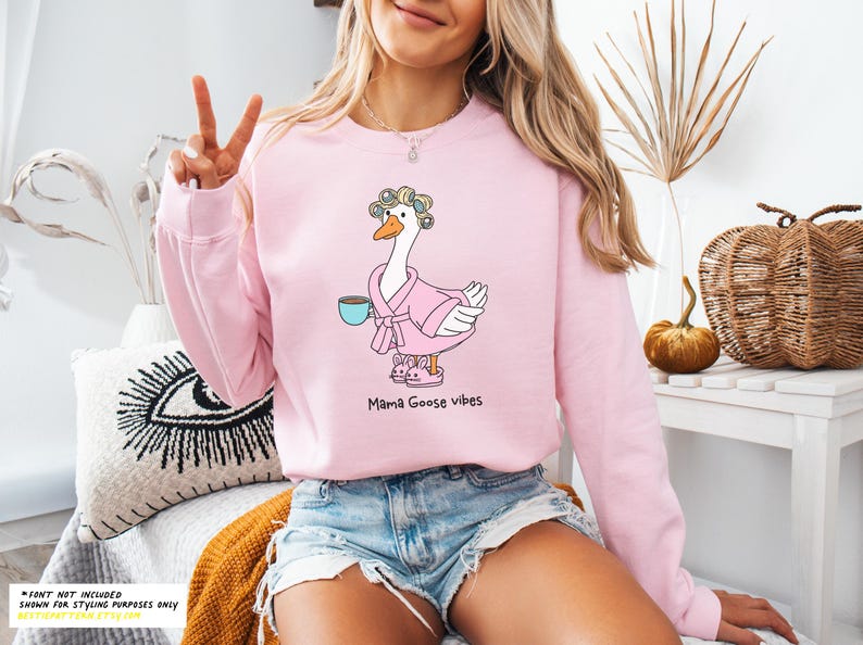 May include: Pink sweatshirt with a cartoon goose wearing a pink robe and holding a cup of coffee. The goose has a hair roller in its hair. The text "Mama Goose Vibes" is printed below the goose.