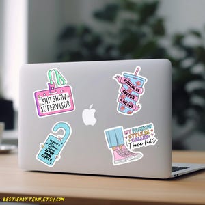 May include: Four colorful stickers on a silver laptop. The stickers feature humorous sayings about coffee, fashion, and motherhood. The stickers are pink, blue, and green with white text.