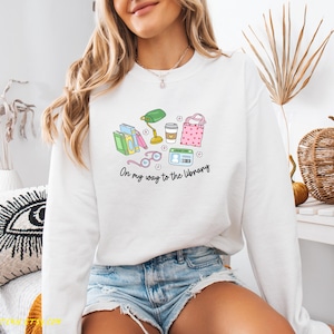 May include: White sweatshirt with a colorful graphic of books, glasses, a lamp, coffee, and a tote bag. The text "On my way to the library" is printed below the graphic. The sweatshirt is worn by a person.