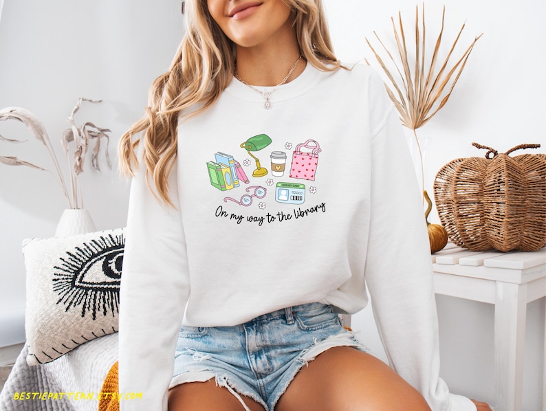May include: White sweatshirt with a colorful graphic of books, glasses, a lamp, coffee, and a tote bag. The text "On my way to the library" is printed below the graphic. The sweatshirt is worn by a person.