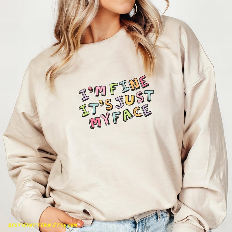 May include: A beige sweatshirt with the text "I'm fine it's just my face" in colorful letters.
