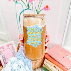 May include: A clear glass tumbler with a wooden lid and a glass straw, filled with a coffee drink. A sticker on the glass reads "Oops I borrowed more library books." Several books are visible in the background.