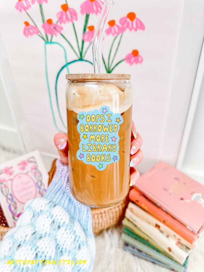 May include: A clear glass tumbler with a wooden lid and a glass straw, filled with a coffee drink. A sticker on the glass reads "Oops I borrowed more library books." Several books are visible in the background.