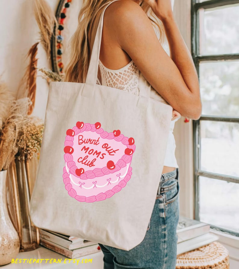 May include: A white canvas tote bag with a pink heart-shaped cake design. The cake has red cherries and the text "Burnt out Moms Club".