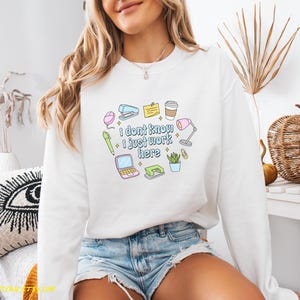 May include: White sweatshirt with the text "I don't know I just work here" and colorful office supply graphics. The design includes a stapler, coffee cup, laptop, and a desk lamp.