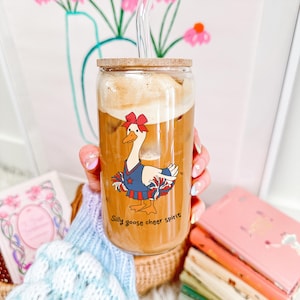 May include: A glass with a wooden lid and a clear straw filled with a brown beverage. The glass has a cartoon image of a goose wearing a blue and red cheerleading outfit with the text "Silly goose cheer spirit".