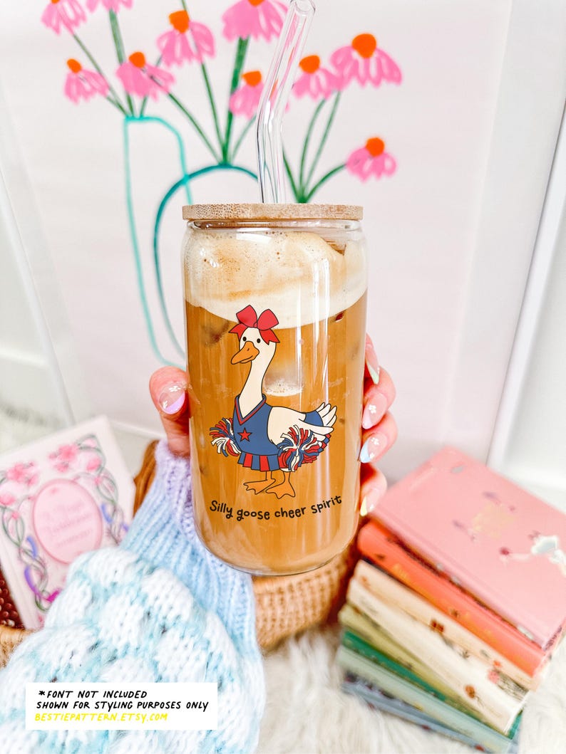 May include: A glass with a wooden lid and a clear straw filled with a brown beverage. The glass has a cartoon image of a goose wearing a blue and red cheerleading outfit with the text "Silly goose cheer spirit".