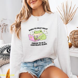 Puede incluir: Sudadera blanca con una rana de dibujos animados con corbata y trabajando en un port&aacute;til rosa. El texto dice: "Born to dilly dally, forced to be a corporate slave."