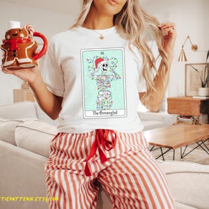May include: White t-shirt with a Christmas-themed tarot card design featuring a skeleton tangled in Christmas lights. The card is titled "The Entangled".