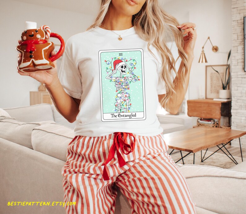 May include: White t-shirt with a Christmas-themed tarot card design featuring a skeleton tangled in Christmas lights. The card is titled "The Entangled".