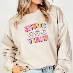 Puede incluir: Una sudadera beige con el texto "JESUS VIBES" en colores arco&iacute;ris y margaritas blancas.