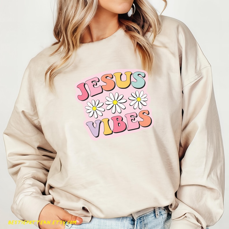 Puede incluir: Una sudadera beige con el texto "JESUS VIBES" en colores arco&iacute;ris y margaritas blancas.