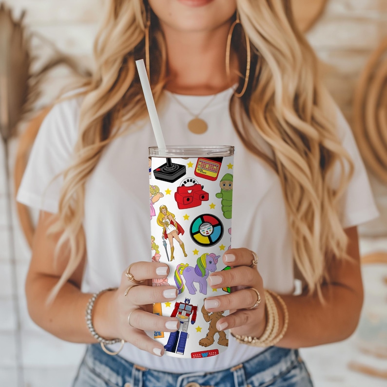 May include: A white stainless steel tumbler with a colourful pattern of 80s toys and characters. The tumbler features a My Little Pony, a Care Bear, a Cabbage Patch Kid, a He-Man figure, a Transformers robot, a Rubik's Cube, a View-Master, a Pac-Man game, and a vintage phone.