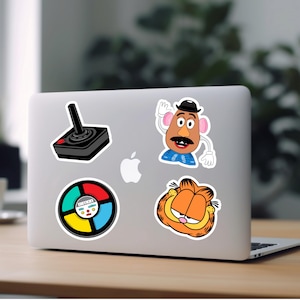 Puede incluir: Un ordenador port&aacute;til con cinco pegatinas. Las pegatinas son de un joystick, una cabeza de patata de dibujos animados, una rueda de juego y un gato de dibujos animados.
