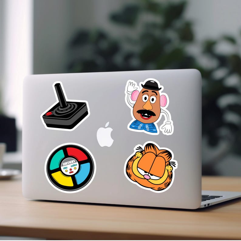 Puede incluir: Un ordenador port&aacute;til con cinco pegatinas. Las pegatinas son de un joystick, una cabeza de patata de dibujos animados, una rueda de juego y un gato de dibujos animados.