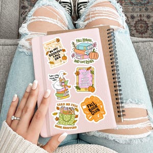May include: A pink notebook with fall-themed stickers, including pumpkins and text like "Fall Leaves" and "Bookish Fall Era." The notebook is held on a person's lap, with distressed jeans and black sneakers visible.