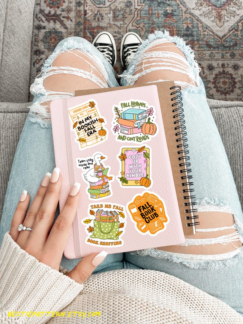 May include: A pink notebook with fall-themed stickers, including pumpkins and text like "Fall Leaves" and "Bookish Fall Era." The notebook is held on a person's lap, with distressed jeans and black sneakers visible.