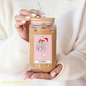 May include: A glass mason jar with a wooden lid and a straw filled with iced coffee. A pink and white tarot card with a skeleton holding presents and the text "The Gifter" is adhered to the front of the jar.