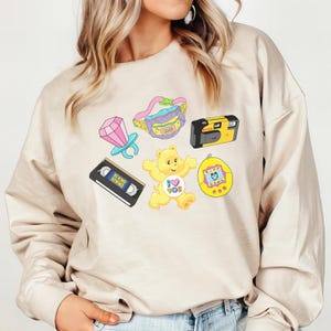 May include: A beige sweatshirt with a graphic print of a Care Bear, a cassette tape, a camera, a ring, a fanny pack, and a Tamagotchi. The graphic is in a cartoon style with bright colors.