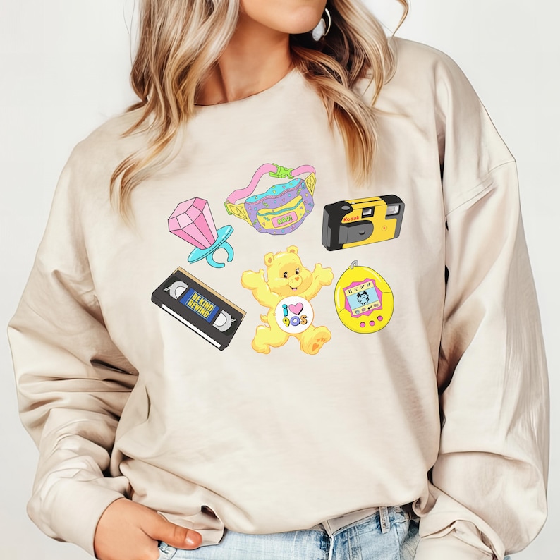 May include: A beige sweatshirt with a graphic print of a Care Bear, a cassette tape, a camera, a ring, a fanny pack, and a Tamagotchi. The graphic is in a cartoon style with bright colors.