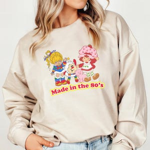May include: A beige sweatshirt featuring a graphic print of three cartoon characters. The characters are a girl with blonde hair, a girl with red hair and a strawberry on her head, and a white fluffy creature. The text "Made in the 80's" is printed below the characters.