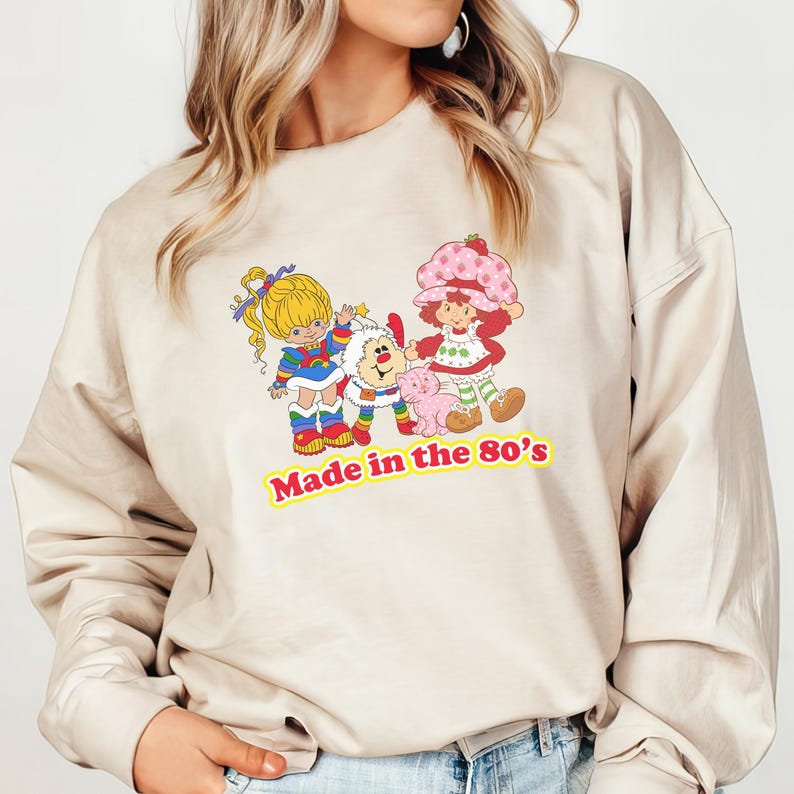 May include: A beige sweatshirt featuring a graphic print of three cartoon characters. The characters are a girl with blonde hair, a girl with red hair and a strawberry on her head, and a white fluffy creature. The text "Made in the 80's" is printed below the characters.